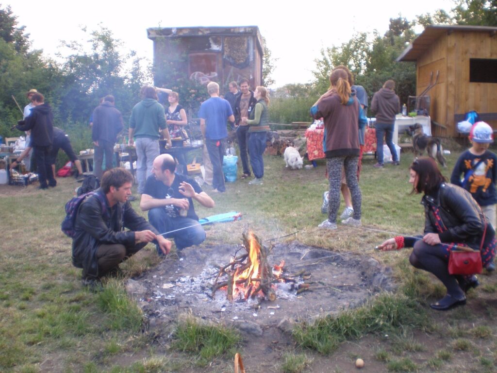 Sousedé z osady Malina stojí a sedí kolem ohně za podvečerního světla
